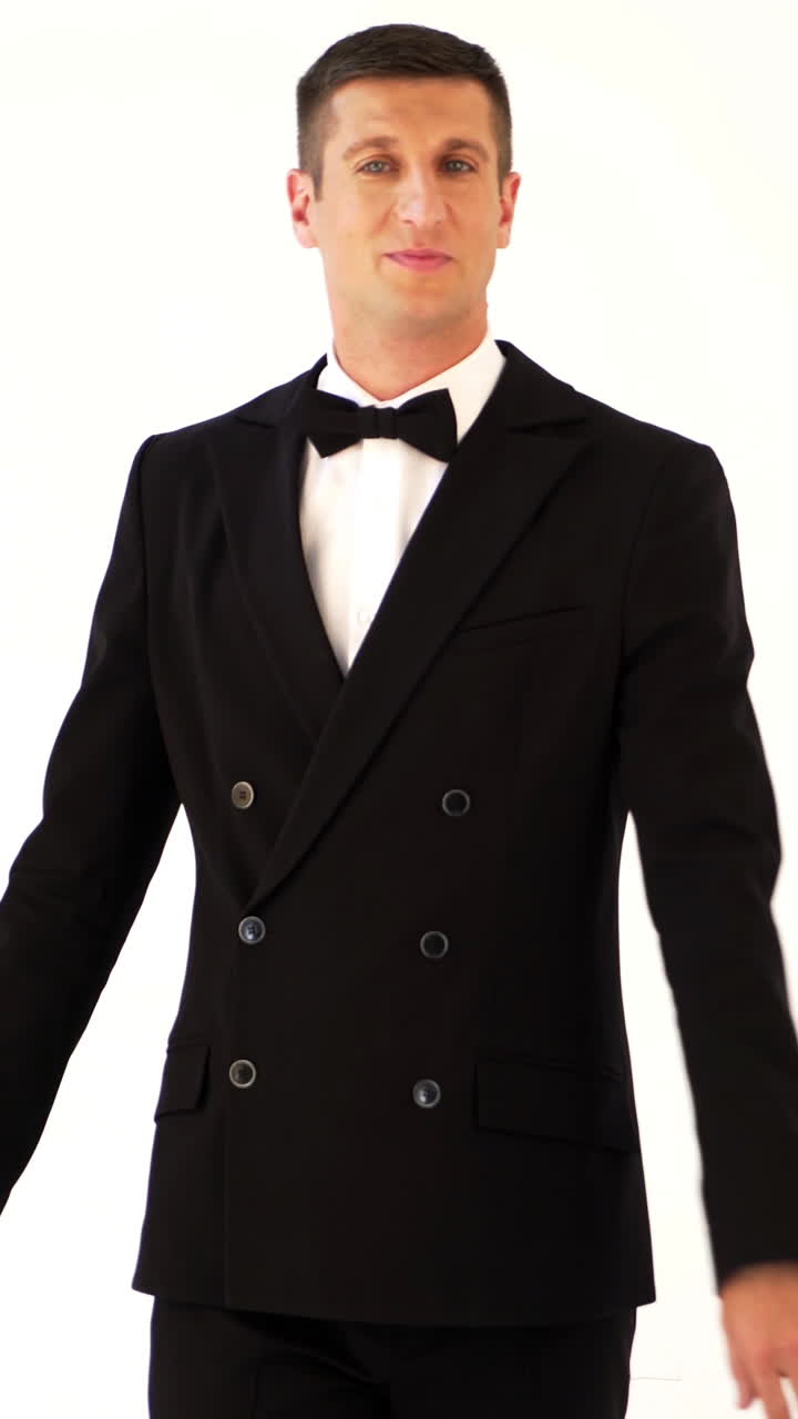 handsome man in a suit with bow-tie is posing in front of a mirror at the exit of the room. Parody of the gentleman on the white background Vertical video