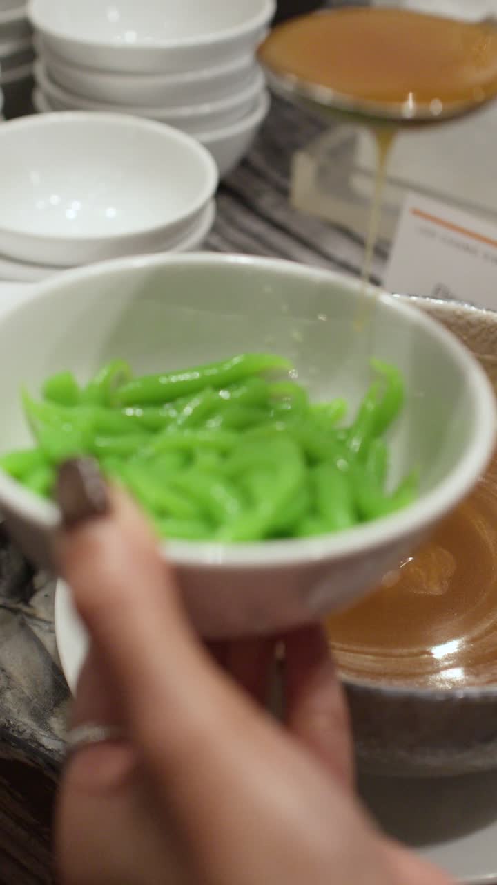 Close-up shot of pouring palm sugar coconut milk over green pandan noodles in a bowl