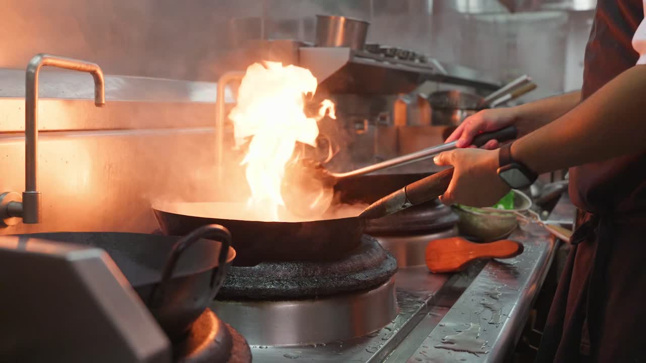 Uno chef pulisce un wok caldo con acqua e vapore usando il suo mestolo sopra le fiamme.