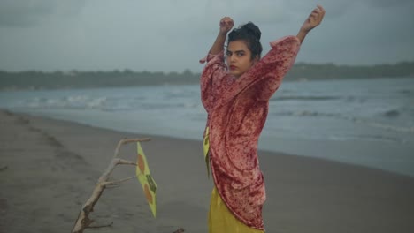 slow motion shot of a woman in a golden dress and red cardigan standing on the beach with her hands in the air