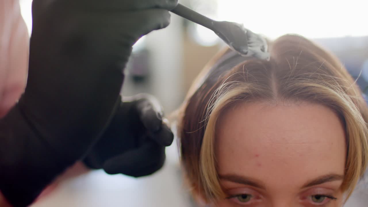 Gloved hand of caucasian hairdresser dyeing female client's hair with brush at salon, in slow motion
