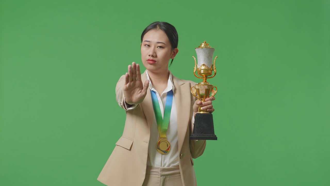 금메달과 트로피를 입은 슈트를 입은 아시아 사업가 여성은 손 표지 없이 불쾌감을 표하며 스튜디오의 녹색 화면 배경에서 카메라에 미소 짓고 있습니다.