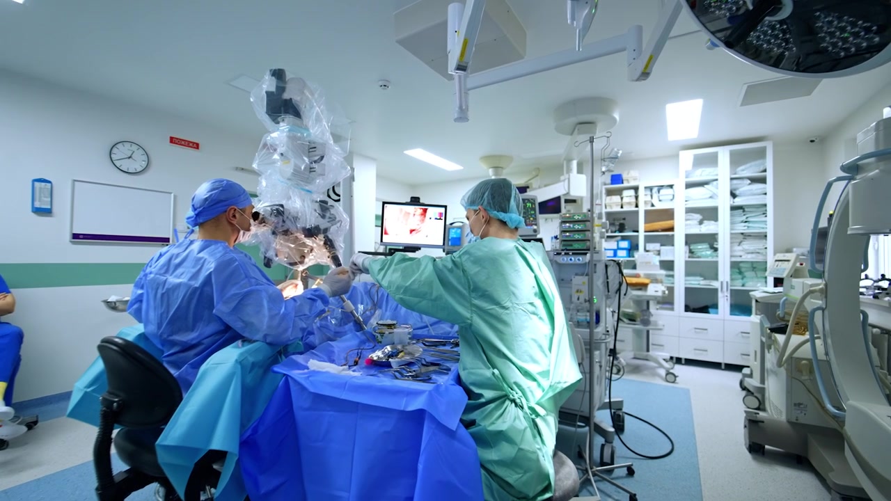 Surgeon looking at microscope performs neurosurgical operation. Assistant sits at the surgical stand preparing tools for the doctor.