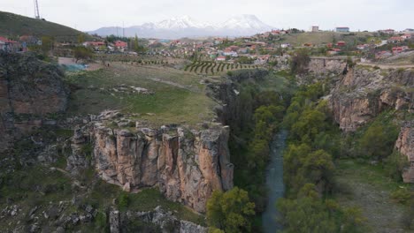 Toma-Aérea-De-Un-Pueblo-En-Los-Acantilados-Del-Cañón-Ihlara-En-Capadocia,-Turquía,-Con-La-Vista-De-Un-Hermoso-Pico-Cubierto-De-Nieve-En-El-Fondo