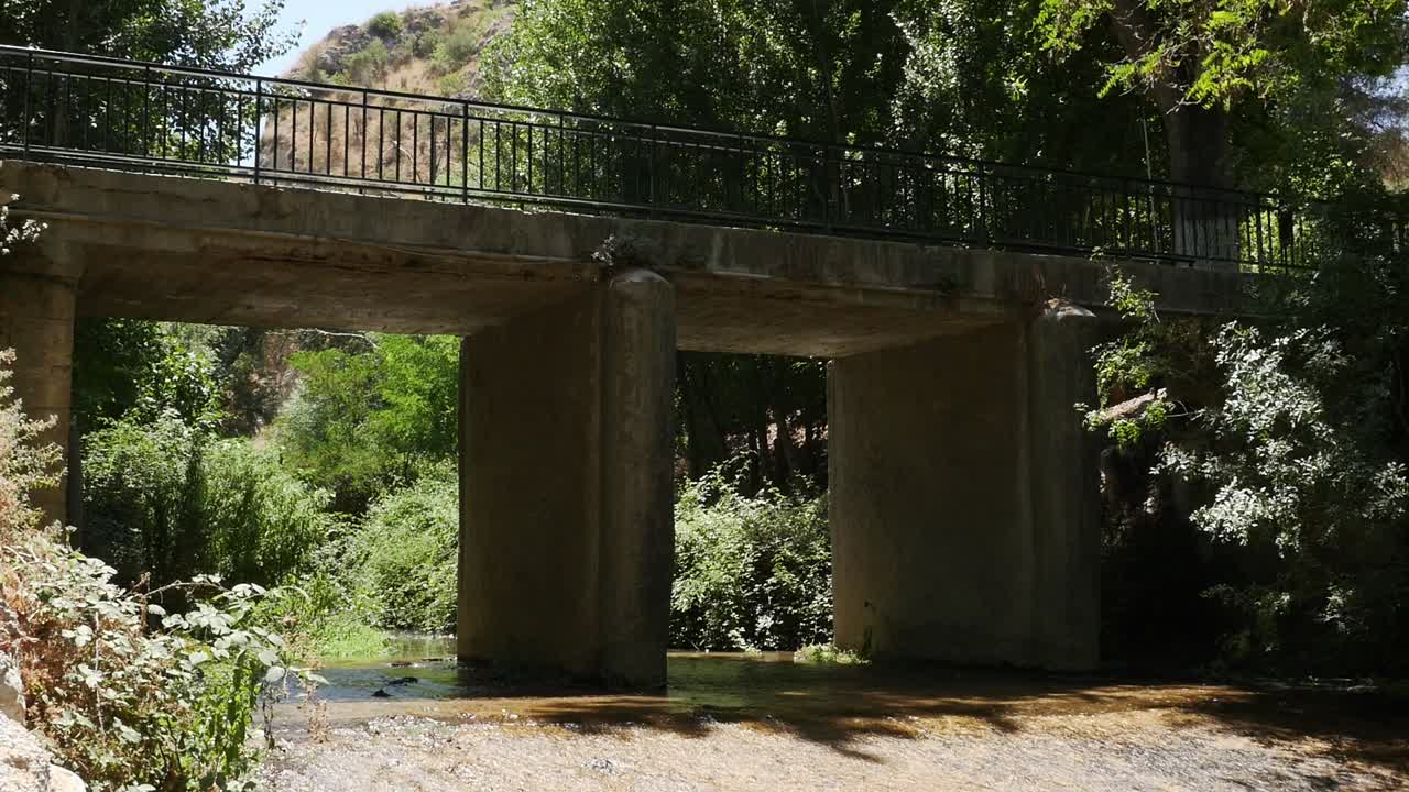 Puente de hormigón con barandilla de hierro que cruza un río tranquilo rodeado de árboles y vegetación en Alhama de Granada
