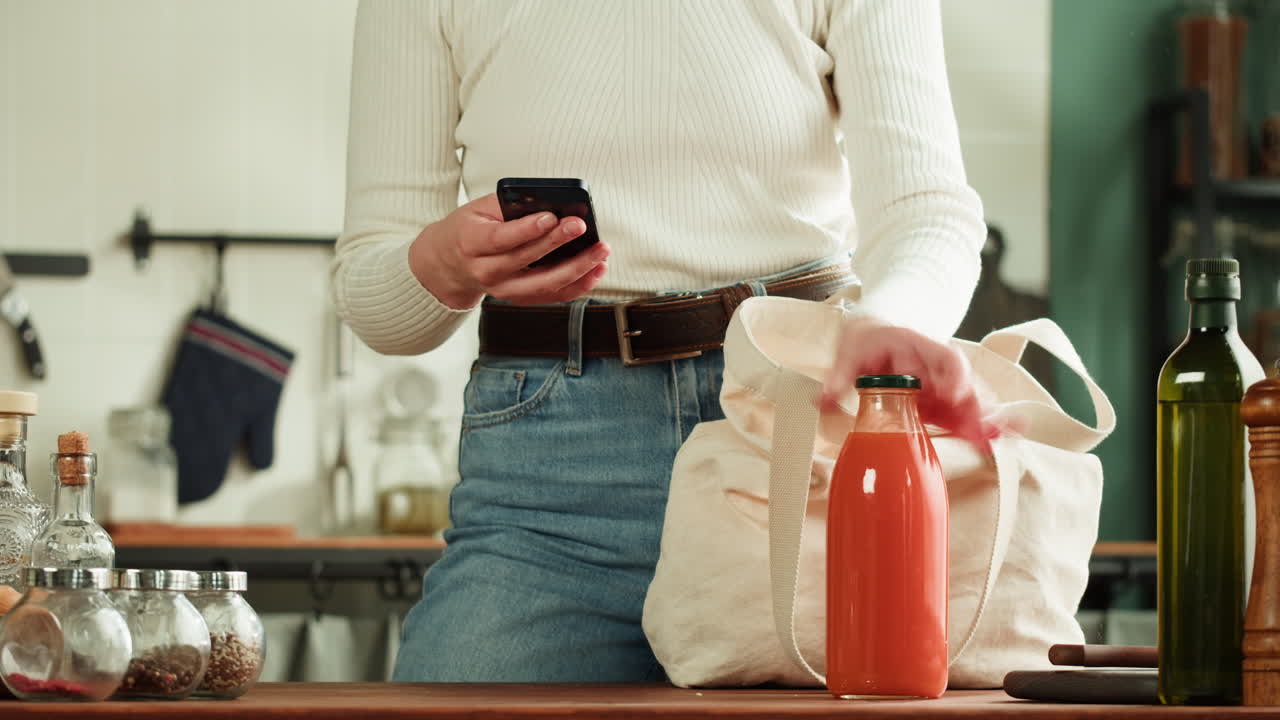 Woman shopping online for groceries, using phone in kitchen