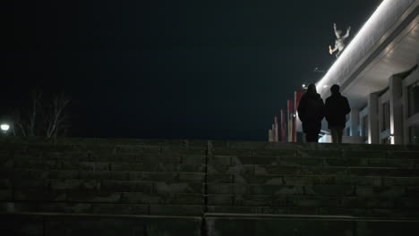 nighttime view of two people walking up a wide, illuminated urban staircase, with a building having light on the linter level