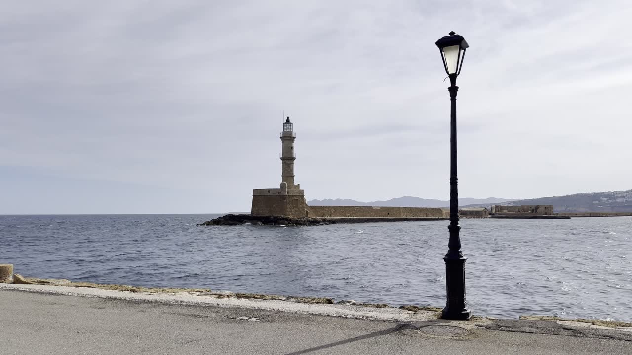Inquadratura cinematografica lenta del faro veneziano a Chania, Creta, con mare calmo, luce soffusa del mattino e lampione tradizionale in primo piano, in una giornata tranquilla e senza persone.