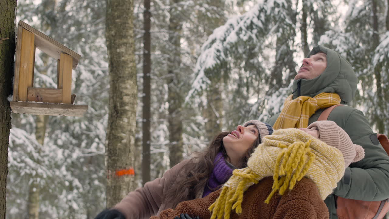 겨울옷을 입은 아버지, 어머니, 딸이 눈 덮인 숲을 올려다보는 나무 앞에 가까이 있습니다.