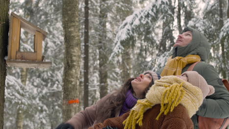 close up view of father, mother and daughter dressed in winter clothes are in front of a tree looking up in a snowy forest