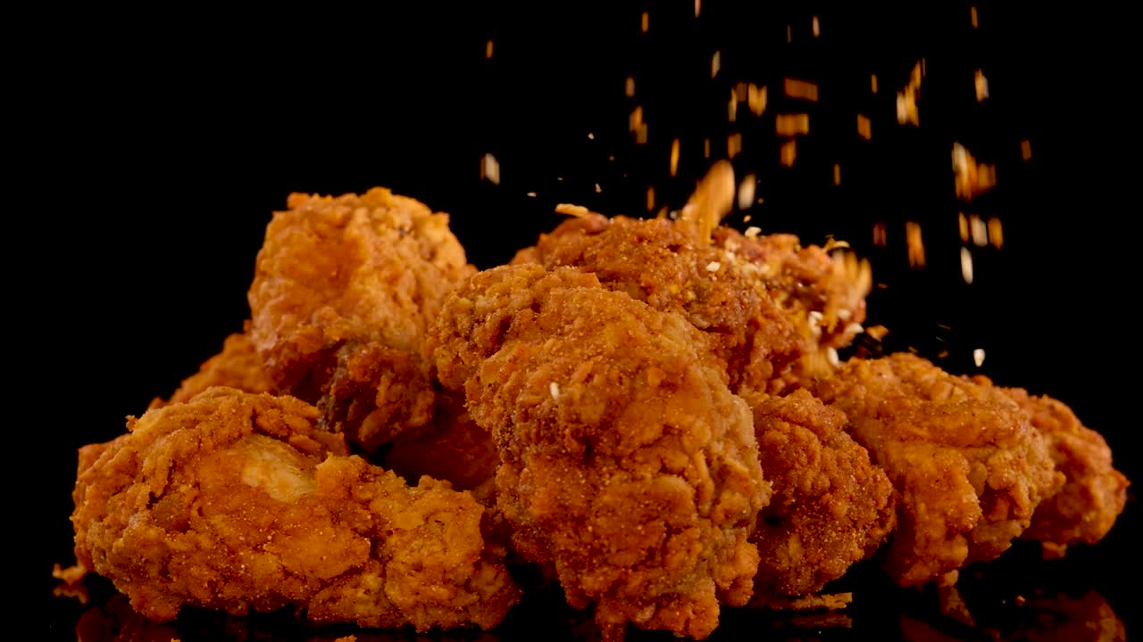 Golden fried chicken pieces are sprinkled with spices in slow motion against a black background, highlighting texture, seasoning, and dramatic studio lighting