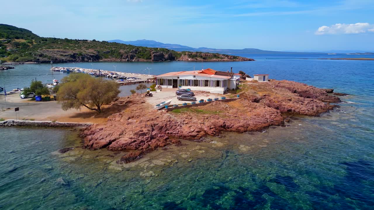 Drone shot of a beach restaurant in Greece, with crystal clear water, people swimming, enjoying boats, and a small port.