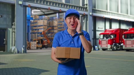 asian male courier in blue uniform smiling and talking to camera while delivering a carton, outside of logistics distributions warehouse