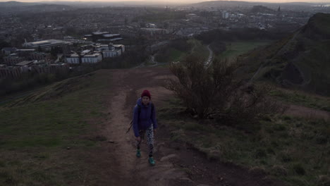 Dolly-shot-of-girl-walking-up-the-Arthurs-seat-mountain-on-the-hiking-trail-in-evening-during-golden-hour-with-city-of-Edinburgh-in-the-background