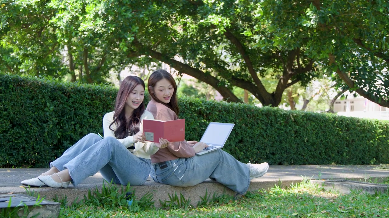 明るい日差しの屋外で、2人の女子学生が背中合わせに座り、ノートパソコンとノートを使っている。