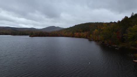 Moody-aerial-view-over-dark-lake-near-colorful-fall-forest-hillside-before-rain