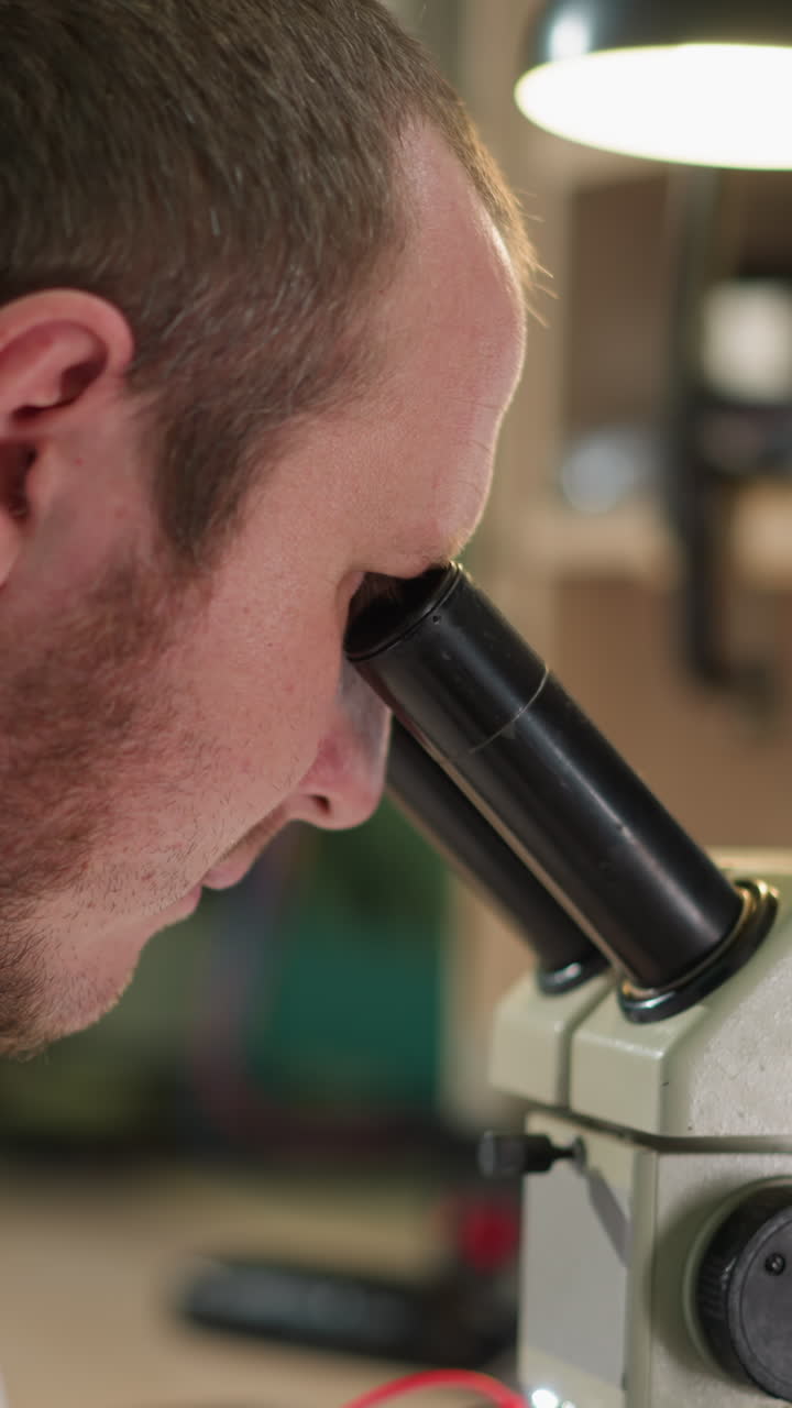A close view of a technician in a white lab coat carefully observing something under a microscope with a red wire in his hand in a laboratory, the workspace has a desk lamp