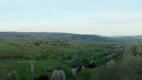 Drone-glides-low-over-blooming-trees-in-Central-Slovakia's-Hrinova-area,-revealing-a-modern-Catholic-church-atop-a-hill