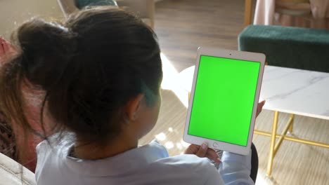 punto de vista de una mujer india que usa una pantalla de maqueta verde, una tableta digital en modo paisaje mientras está sentada en un sofá. en el fondo, una acogedora sala de estar