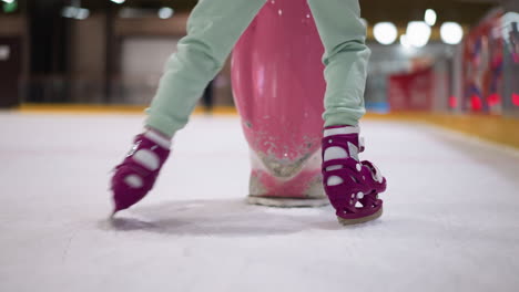 close view of a person wearing light green cloth and purple skates, pushing a pink object on an ice rink