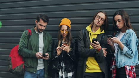 group of caucasian friends leaning on the wall while talking and typing on their smartphones in the street