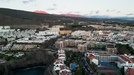 el dron captura la ciudad de los gigantes con su pintoresco encanto costero, mientras que en la distancia, el majestuoso volcán teide de tonos rojos se erige como un punto de referencia icónico en la isla de tenerife.