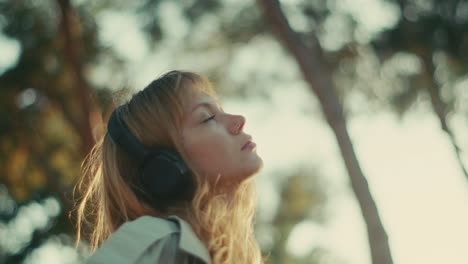 Lady-with-headphones-listens-to-music-at-sunset,-light-breeze-through-loose-hair