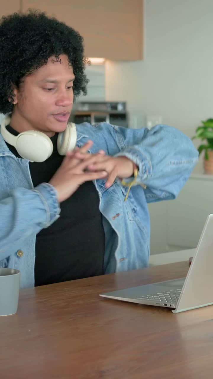 Vertical video: Typing man in denim jacket, headphones, laptop in kitchen pausing checking watch