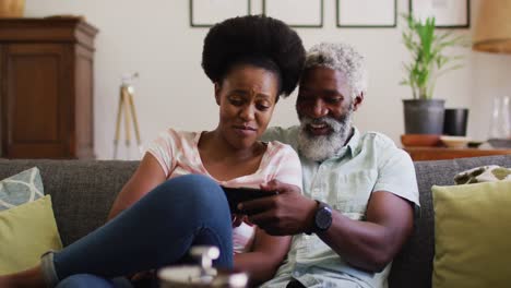 happy african american couple sitting on couch looking at smartphone and smiling