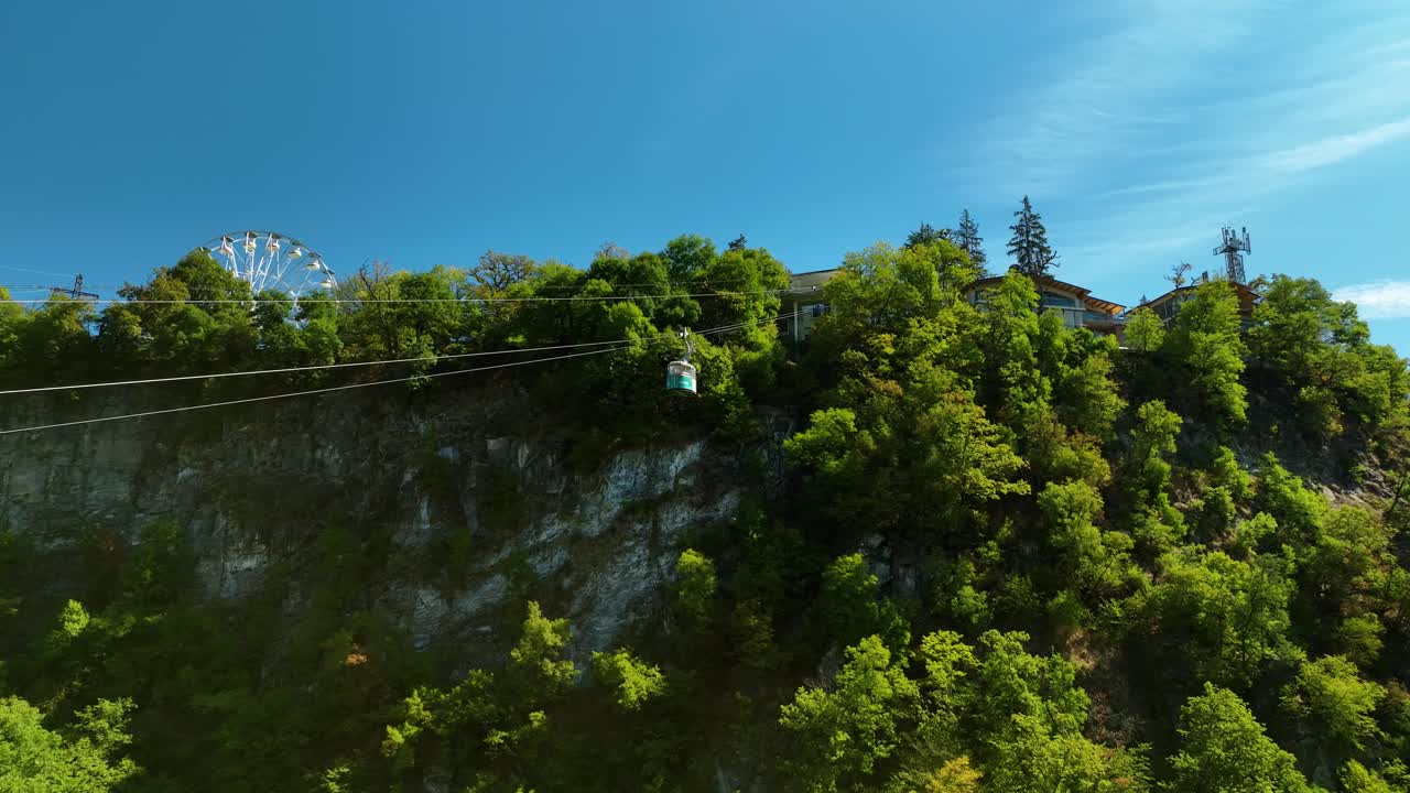 A cable car glides across a high mountain ridge, with lush green trees, rocky cliffs, and a ferris wheel faintly visible in the distance