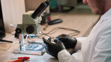 vista de cerca de un técnico en una bata de laboratorio ajustando un tornillo en su mano con un componente electrónico bajo un microscopio y varias herramientas en la mesa y otras cosas en el estante