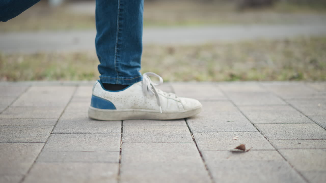 Black guy wearing jeans and white sneakers walking outdoors on paved path during early spring, close-up view of foot in step motion, blurred grass and pathway background, casual urban style in natural daylight