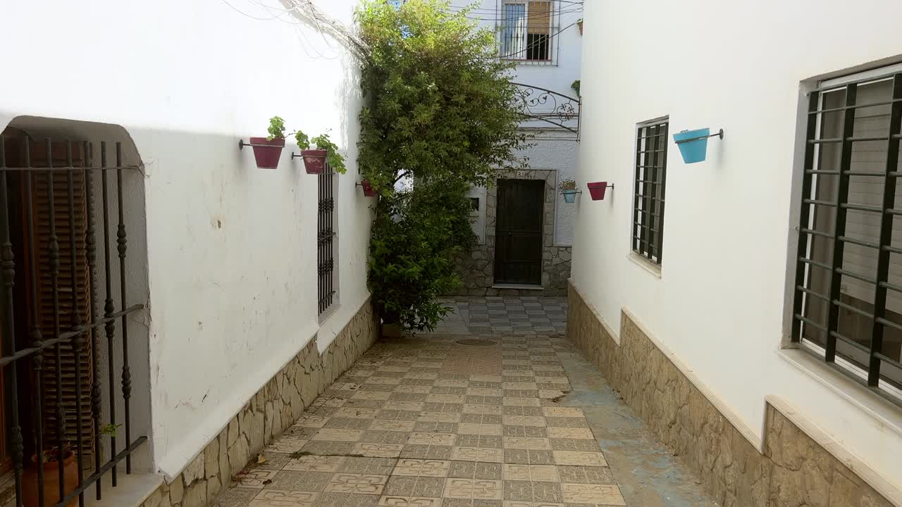 Quiet narrow alley between white houses with colorful flower pots and trees under a bright sky