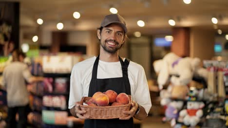 Retrato-De-Un-Feliz-Trabajador-De-Una-Tienda-Con-Una-Camiseta-Blanca-Y-Un-Delantal-Negro,-Un-Hombre-Sosteniendo-Una-Canasta-De-Melocotones-En-Sus-Manos-Y-Sonriendo