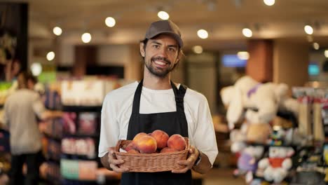 retrato de un trabajador de una tienda feliz en una camiseta blanca y un delantal negro, un hombre sosteniendo una canasta de melocotones en sus manos y sonriendo
