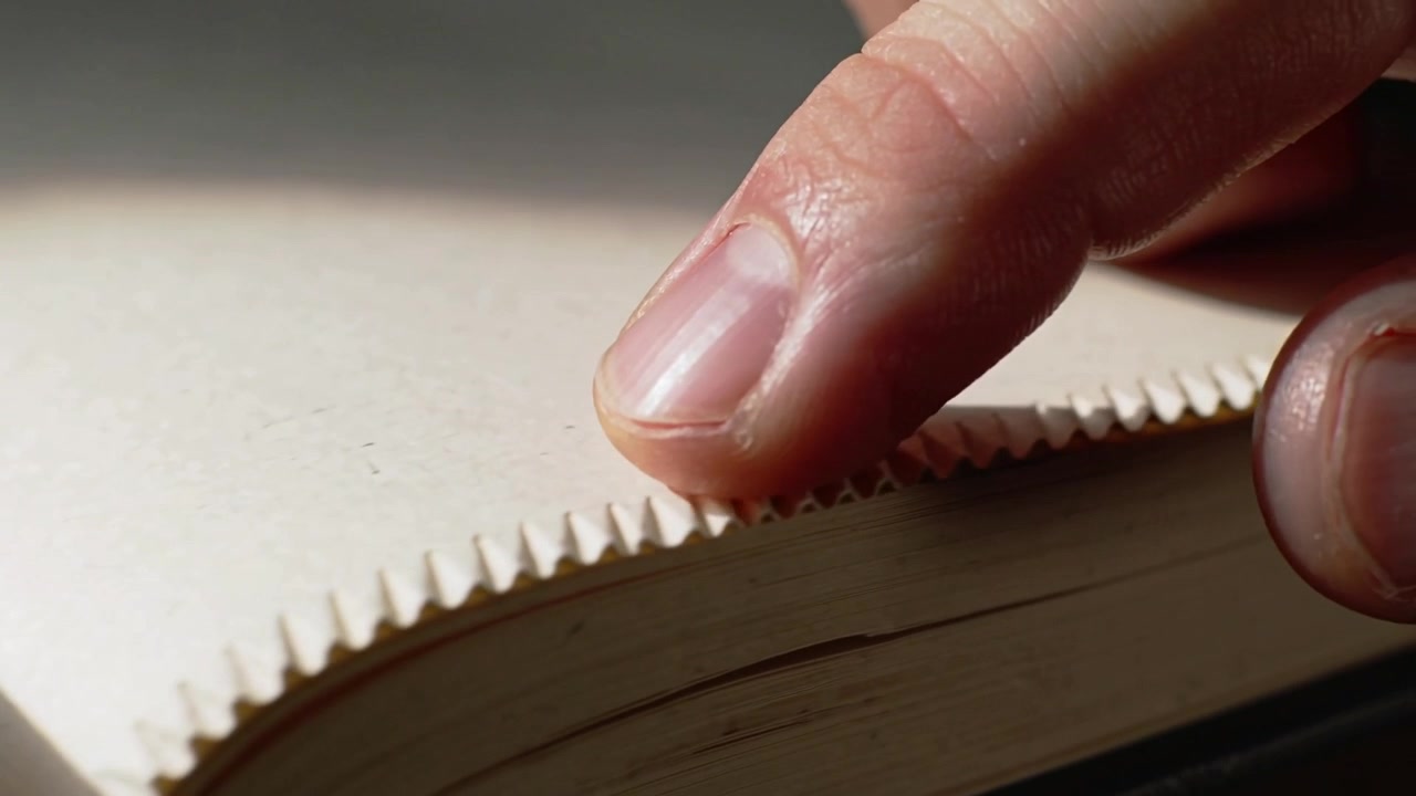 Dito che scorre sul taglio dentellato di un libro sulla scrivania, percependo la texture, il nastro e lo spazio disponibile.