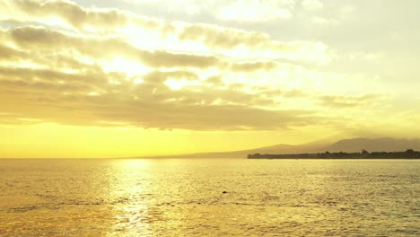 golden sky with glowing clouds hiding sun setting down, reflecting on glassy sea surface around silhouette of tropical island, malaysia