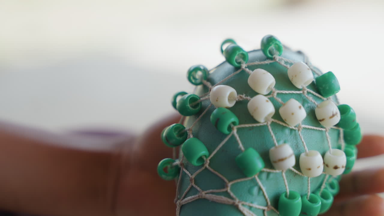 Inspection de perles artisanales. Des mains noires examinent des perles tressées avec soin à la lumière du jour. Vue en gros plan d'un hochet vert orné de perles avec un maillage texturé et une inspection manuelle sous une douce lumière naturelle.