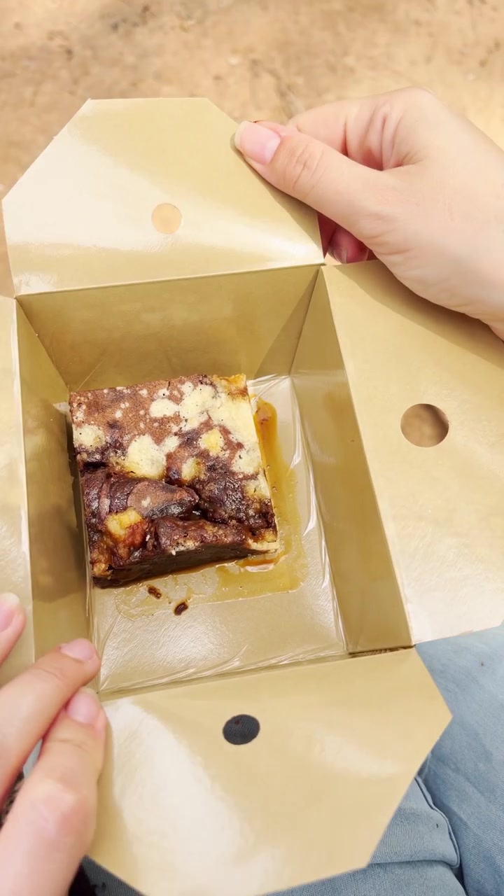 POV mulher sentada no banco desembalando um doce de chocolate dentro de uma caixa de papelão. Embalagem sustentável. Brownie vegano sem glúten. Comendo ao ar livre em um parque público. Reel de vídeo vertical de telefone estilo POV UGC.