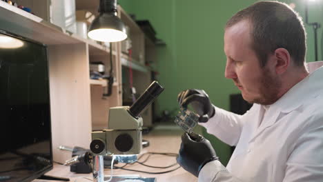 una vista de cerca de un técnico en una bata de laboratorio blanca y guantes negros examinando una placa de circuito bajo un microscopio en un laboratorio de electrónica bien iluminado y otra herramienta en el estante y en la mesa