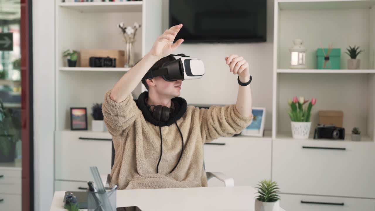 The young man wearing VR goggles is playing a video game and talking. He is sitting at a table, but his hands are actively gesturing in the game.