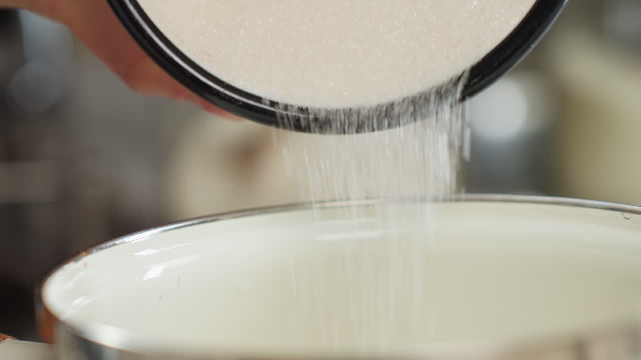 Close up of flour being poured from black-rimmed enamel cup into ceramic bowl during kitchen preparation, with soft background blur suggesting home cooking environment