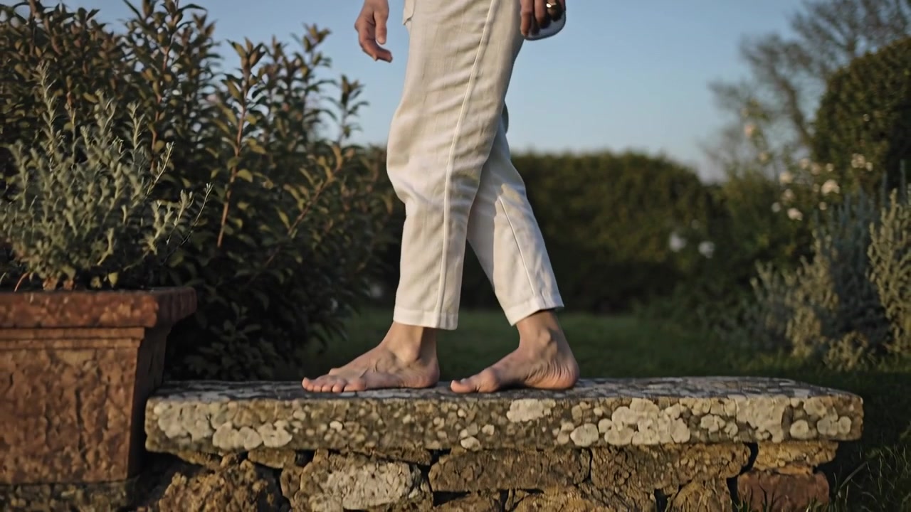 Camera pulling back walking senior in white inspecting plants in dusk garden barefoot on stone wall