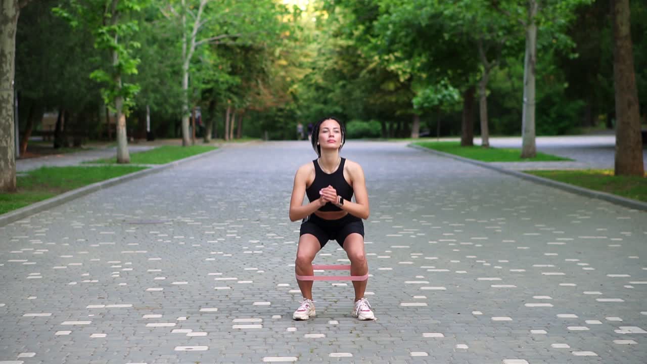 joven atleta hace ejercicios con banda de resistencia haciendo sentadillas al aire libre en el parque público local, verde. mujer atractiva vistiendo