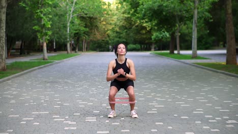 Junge-Sportlerin-Trainiert-Mit-Widerstandsband-Und-Macht-Kniebeugen-Im-Freien-Im-örtlichen,-Grünen-öffentlichen-Park.-Attraktive-Frau-Trägt