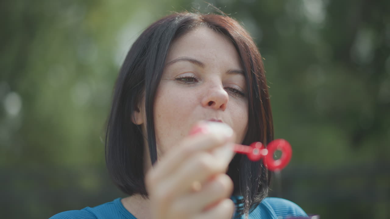 Eine Frau, die draußen Seifenblasen macht, nachdenkliches Nahaufnahme-Porträt mit rotem Zauberstab, weiches Bokeh, grüner Park-Hintergrund, sanftes Sommerlicht, Sommersprossen-Hautdetails, langsame, verspielte Bewegung und friedlich
