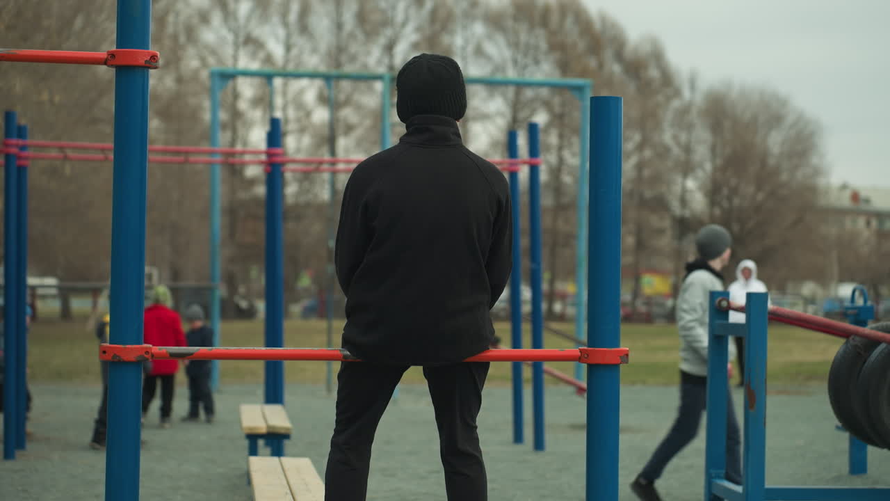 Back view of a person dressed in black and wearing, sitting on a red iron bar in a playground, in the background, children play and another individual in a gray top walks by