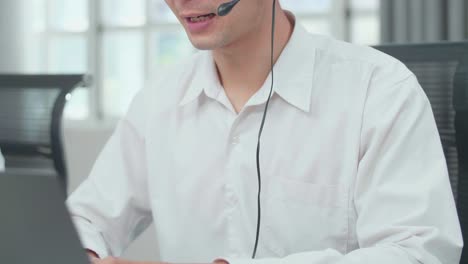 primer plano de un hombre asiático agente de centro de llamadas con auriculares escribiendo en la computadora mientras habla con el cliente en la llamada mientras trabaja con colegas en la oficina