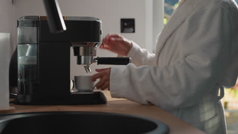 woman puts cup on coffee machine water tray in kitchen closeup. lady in cozy clothes fills mug with hot strong drink from coffeemaker at home. domestic appliances