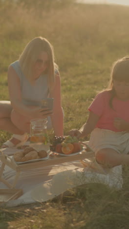 madre e hija sentadas al aire libre compartiendo un picnic relajante bajo la luz del sol, la madre tomando una selfie mientras la hija come frutas frescas. la alfombra de picnic tiene bocadillos, frutas y jarro de agua